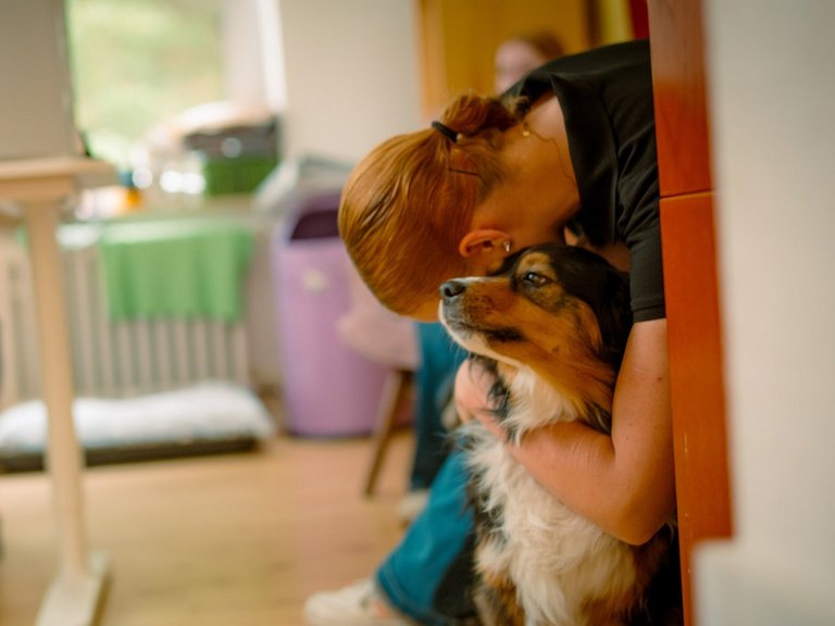 Eine Frau mit roten Haaren lehnt an einer Wand und umarmt zärtlich einen braun-weißen Hund, der zu ihr aufblickt. Im unscharfen Hintergrund ist ein Büroarbeitsplatz zu erkennen.