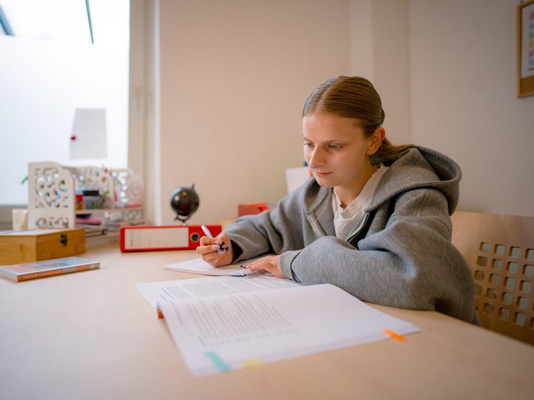 Eine junge Frau in einem grauen Kapuzenpullover sitzt an einem Schreibtisch und schreibt konzentriert auf ein Blatt Papier. Vor ihr liegen weitere Unterlagen.