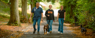 Drei Personen gehen mit zwei Hunden auf einem asphaltierten Waldweg spazieren. Ein Mann in blauem Hemd und zwei Frauen in schwarzen Oberteilen laufen lächelnd auf den Betrachter zu. Der Weg ist von Bäumen gesäumt und mit herbstlichen Blättern bedeckt. Zwei kleine Hunde an der Leine begleiten sie.